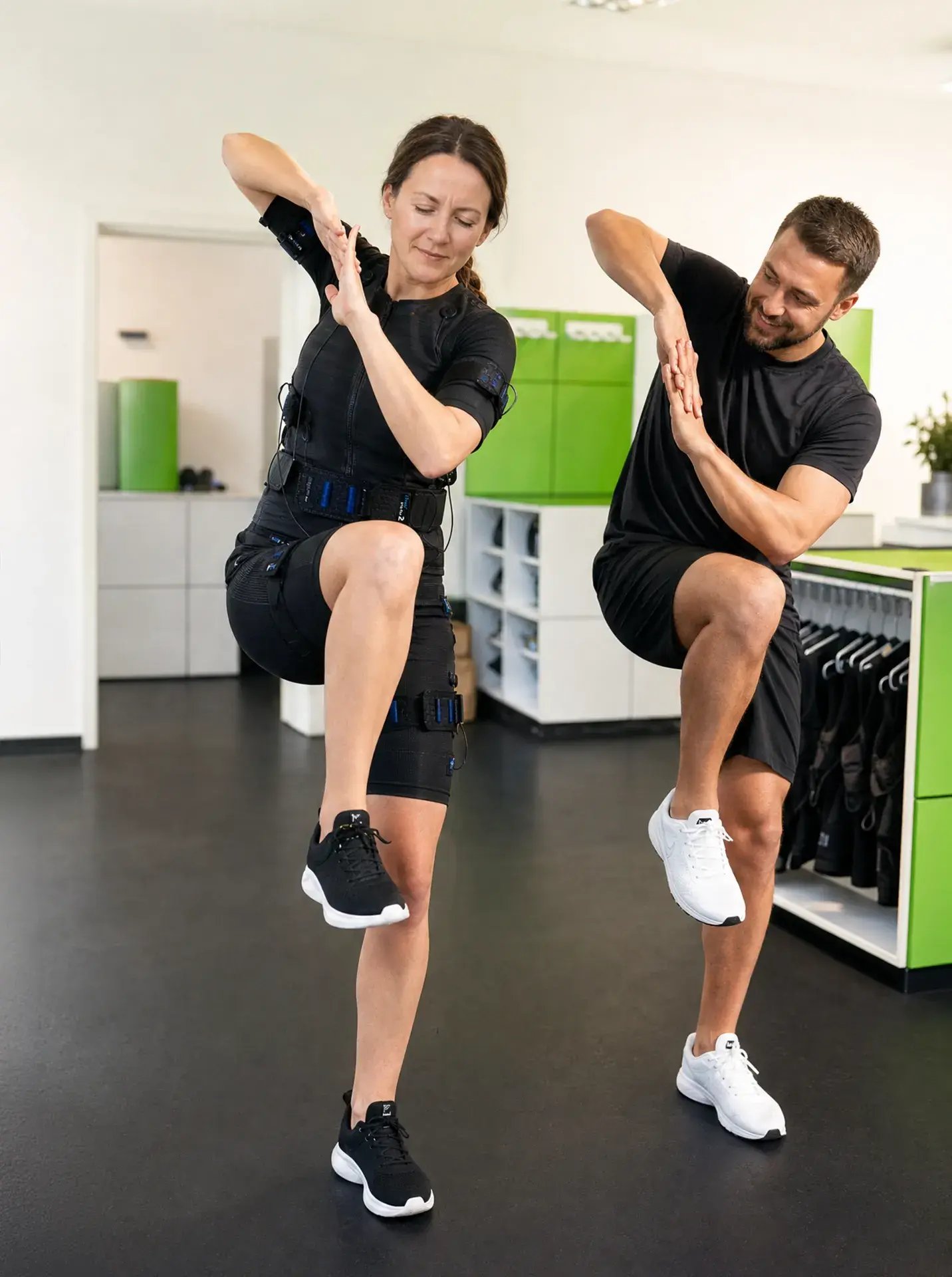 Trainer macht Übung vor, Frau im EMS-Anzug hebt das Knie zur Rumpfstabilisation bei Rückenschmerzen